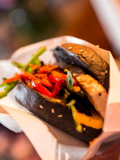 A handheld burger with a black bun, filled with vegetables and a slice of meat. The colorful ingredients create an appetizing appearance. | © Street Food Market Austria