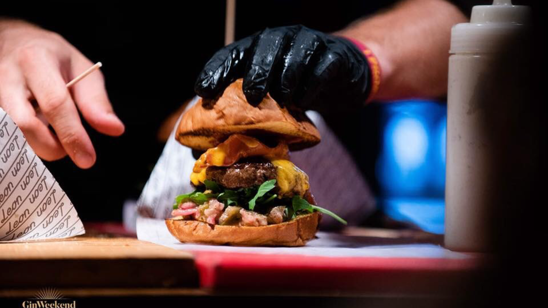 Ein leckerer Hamburger wird mit frischen Zutaten zubereitet. Die Hand mit einem schwarzen Handschuh hält das Brötchen über dem bunten Belag. | © Street Food Market Austria