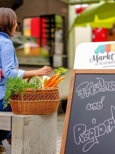 Ein Paar sitzt auf einer Bank in einem Markt, umgeben von frischen Karotten und einem Schild mit der Aufschrift „Frisches und Regionales“. Die Atmosphäre ist lebhaft und einladend. | © Markt´ln Knittelfeld