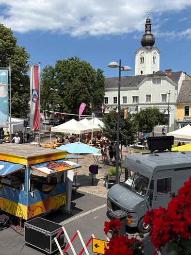 Ein lebendiger Marktplatz mit vielen Food-Trucks und Ständen. Im Hintergrund ist ein schöner Kirchturm und blühende Bäume zu sehen.