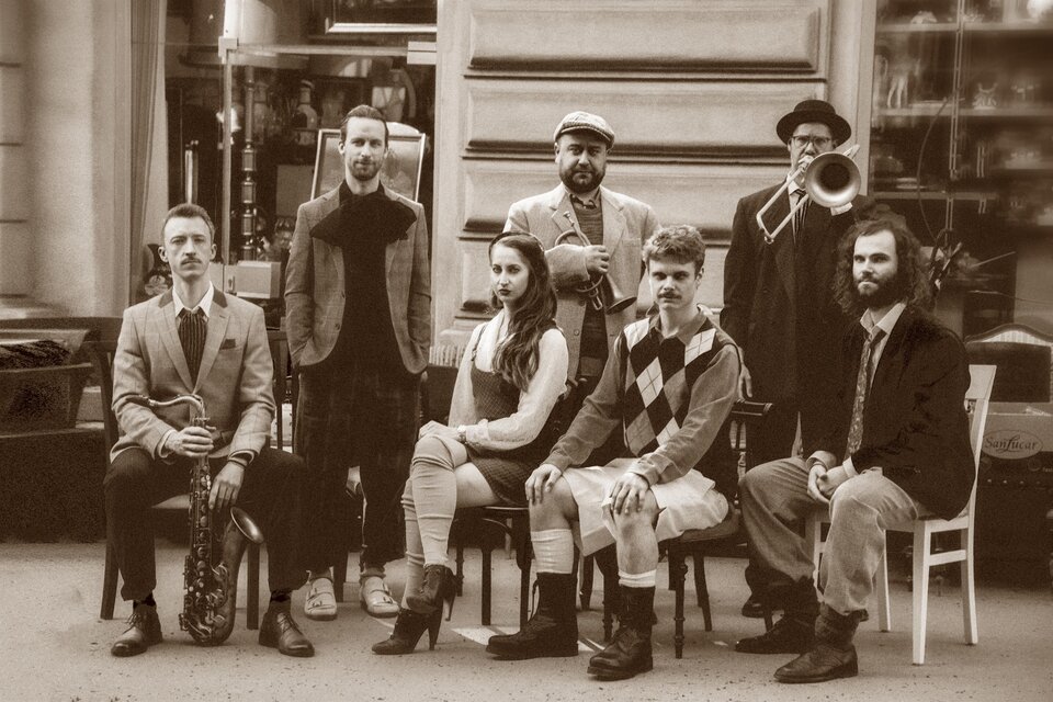 Eine Gruppe von sieben Personen sitzt und steht zusammen, gekleidet in stilvolle vintage Kleidung. Sie scheinen Teil einer Musik- oder Unterhaltungstruppe zu sein, mit Instrumenten in der Nähe. | © Streetviewe Dixieclub