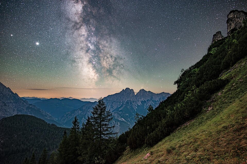 Eine beeindruckende Berglandschaft unter dem Sternenhimmel. Die Milchstraße ist klar sichtbar, umgeben von hohen Bergen und Bäumen. | © Andreas Hollinger