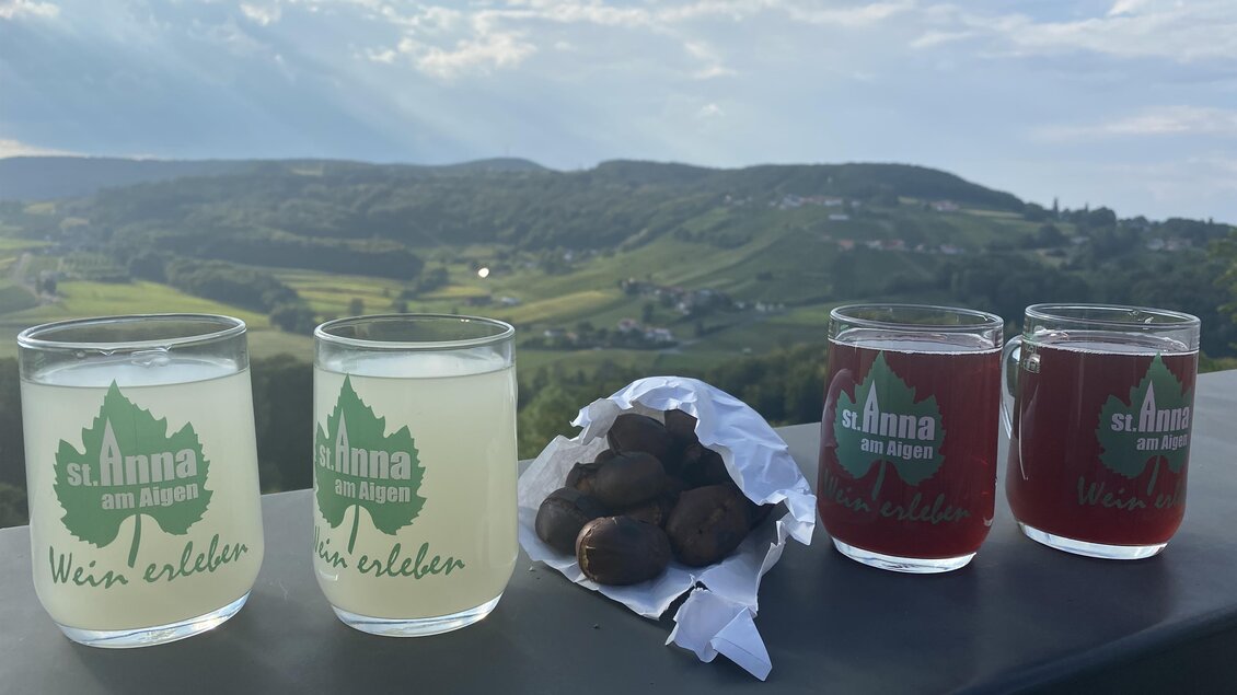Zwei Gläser mit Getränken stehen auf einem Tisch, links ein klares, weißes Getränk und rechts ein rotes Getränk. Neben den Gläsern liegt eine Packung mit Snacks und im Hintergrund erstreckt sich eine grüne Landschaft. | © Karin Grießbacher