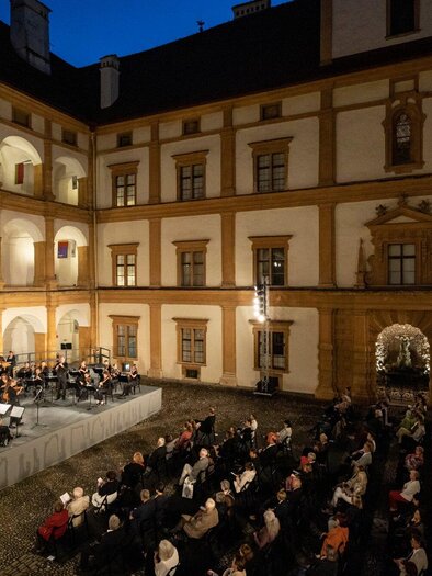 An orchestra is playing on a stage in the courtyard of a historic building.  
The audience is sitting in rows and enjoying the concert at night. | © Nikola Milatovic