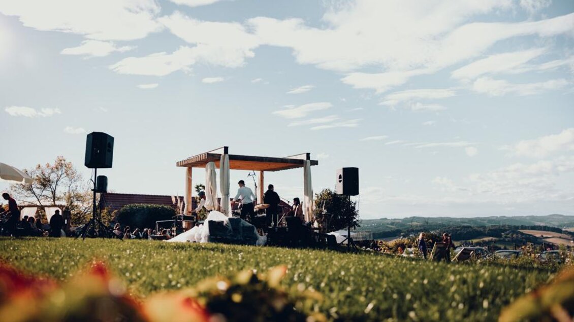 **Musik unter freiem Himmel in traumhafter Kulisse:** Eine Bühne inmitten der sanften Hügellandschaft, entspannte Atmosphäre und begeisterte Besucher machen dieses Open-Air zu einem besonderen Erlebnis beim Obstbau Haas in Poppendorf. | © Obstbau Haas