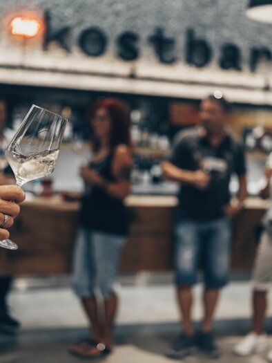 A person is holding a wine glass in their hand, while in the background a bar with various guests can be seen. The atmosphere is relaxed and sociable. | © Ian Ehm