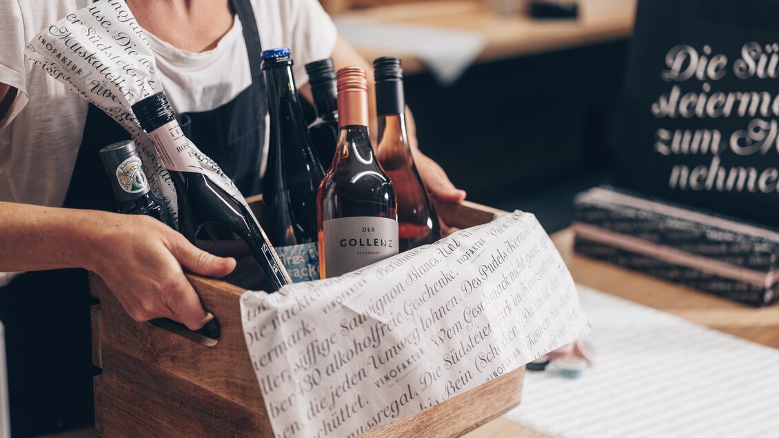 Eine Person hält eine Holzkiste mit verschiedenen Flaschen Wein und Sekt. Im Hintergrund sind dekorative Accessoires zu sehen. | © Ian Ehm