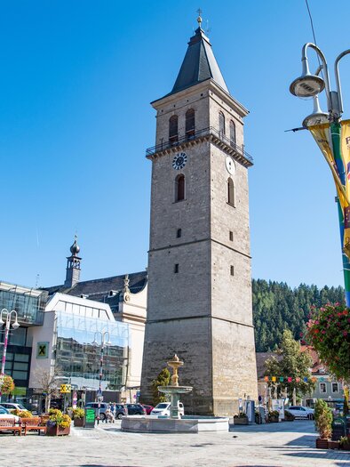 Ein historischer Turm auf einem belebten Platz. Im Hintergrund sind moderne Gebäude und grüne Bäume zu sehen. | © Fössl Anita