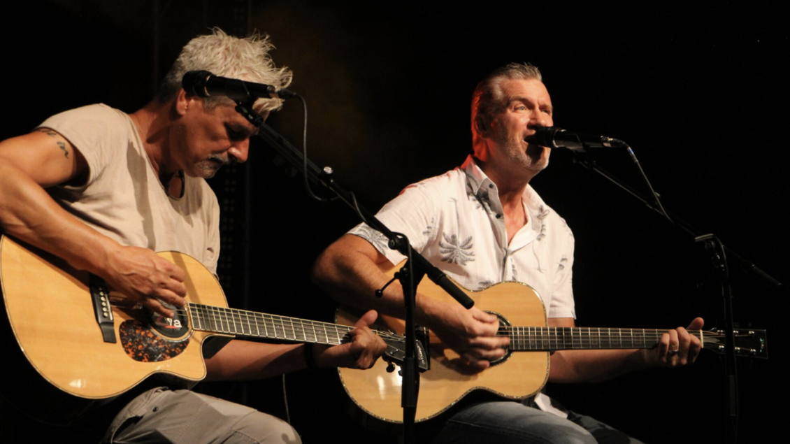 Zwei Musiker sitzen auf der Bühne und spielen Gitarre. Beide singen in ein Mikrofon und sind von Lichtern umgeben. | © Tourismus Oststeiermark