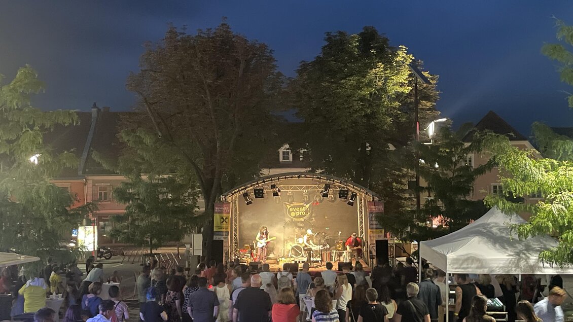 Ein Konzert im Freien bei Nacht mit einer großen Menschenmenge. Die Bühne ist gut beleuchtet und es sind mehrere Musiker zu sehen.