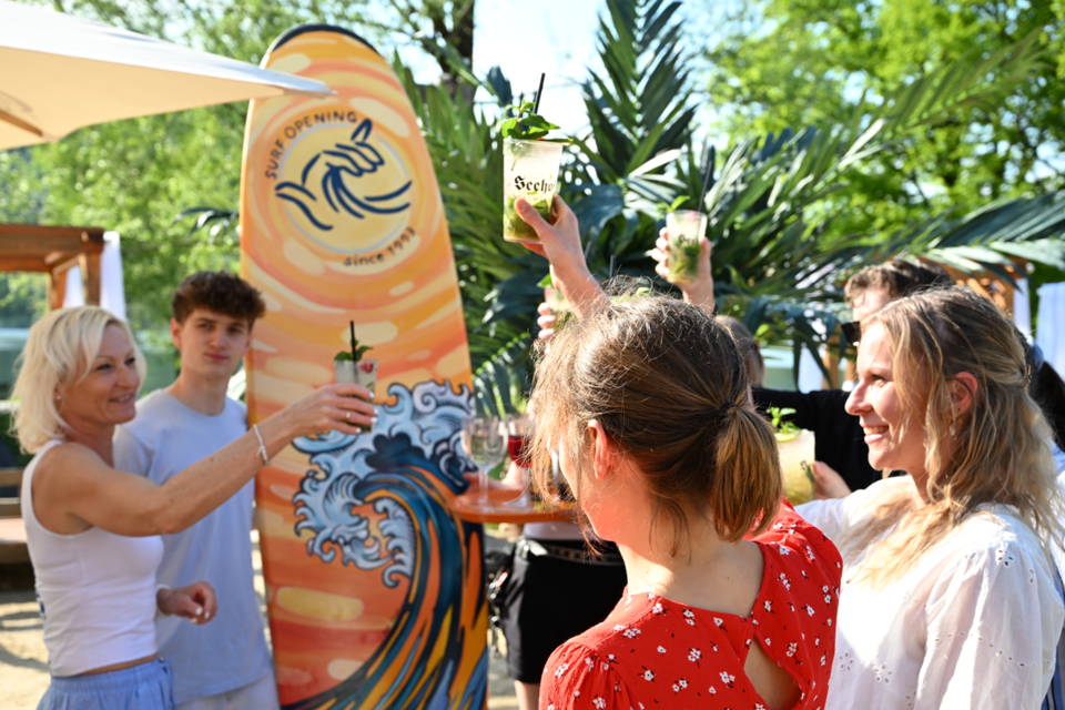 Eine feierliche Atmosphäre mit Freunden, die mit Getränken anstoßen. Im Hintergrund ist eine Surf-Überschrift und grüne Pflanzen zu sehen. | © Oststeiermark Tourismus