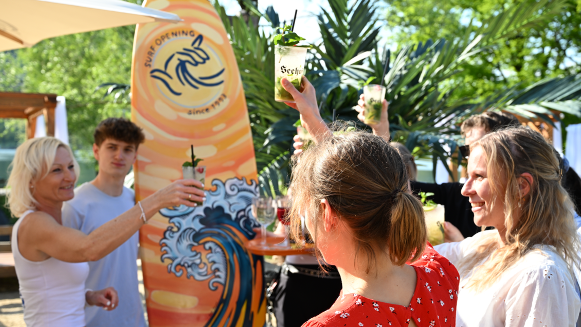 Eine feierliche Atmosphäre mit Freunden, die mit Getränken anstoßen. Im Hintergrund ist eine Surf-Überschrift und grüne Pflanzen zu sehen. | © Oststeiermark Tourismus