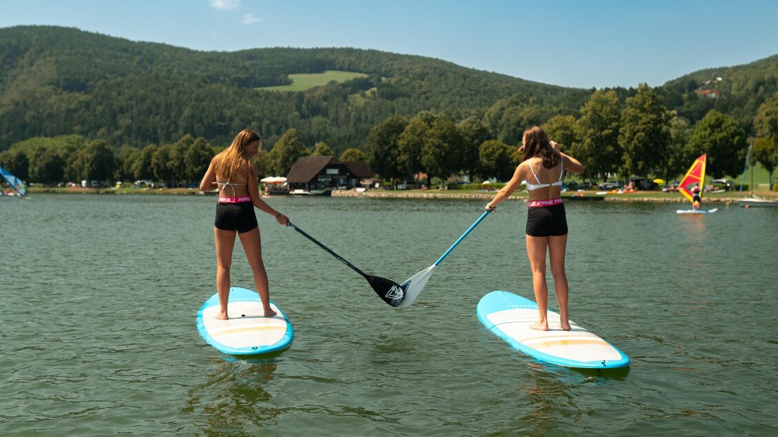 Zwei Mädchen stehen auf Stand-Up-Paddle-Boards im Wasser. Im Hintergrund sind grüne Hügel und Bäume zu sehen. | © Oststeiermark Tourismus