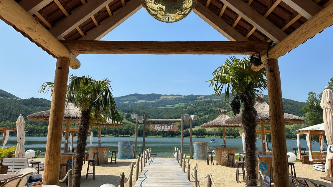 Ein schöner Blick auf einen Strand mit Palmen und einem ruhigen See. Im Hintergrund sind Hütten und grüne Hügel zu sehen. | © Oststeiermark Tourismus