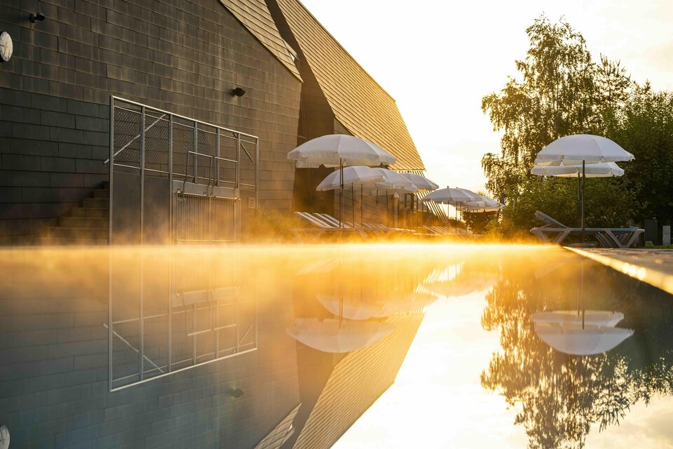 Ein ruhiger Pool mit sanftem Dampf und Sonnenschirmen im Hintergrund. Die warmen Farben des Sonnenuntergangs reflektieren sich im Wasser. | © AQUALUX Therme