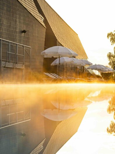 A tranquil pool with gentle steam and umbrellas in the background. The warm colors of the sunset are reflected in the water. | © AQUALUX Therme