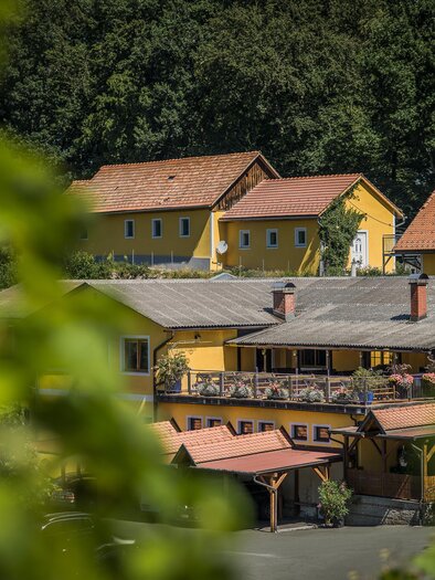 Ein gelbes Gebäude umgeben von Bäumen und Pflanzen. Es gibt eine Terrasse mit Stühlen und Tischen, die einladend wirkt. | © c-mavric