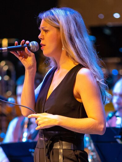 Eine Sängerin steht im Vordergrund und singt in ein Mikrofon. Im Hintergrund sieht man Musiker mit Instrumenten. | © Zauberbilder.at