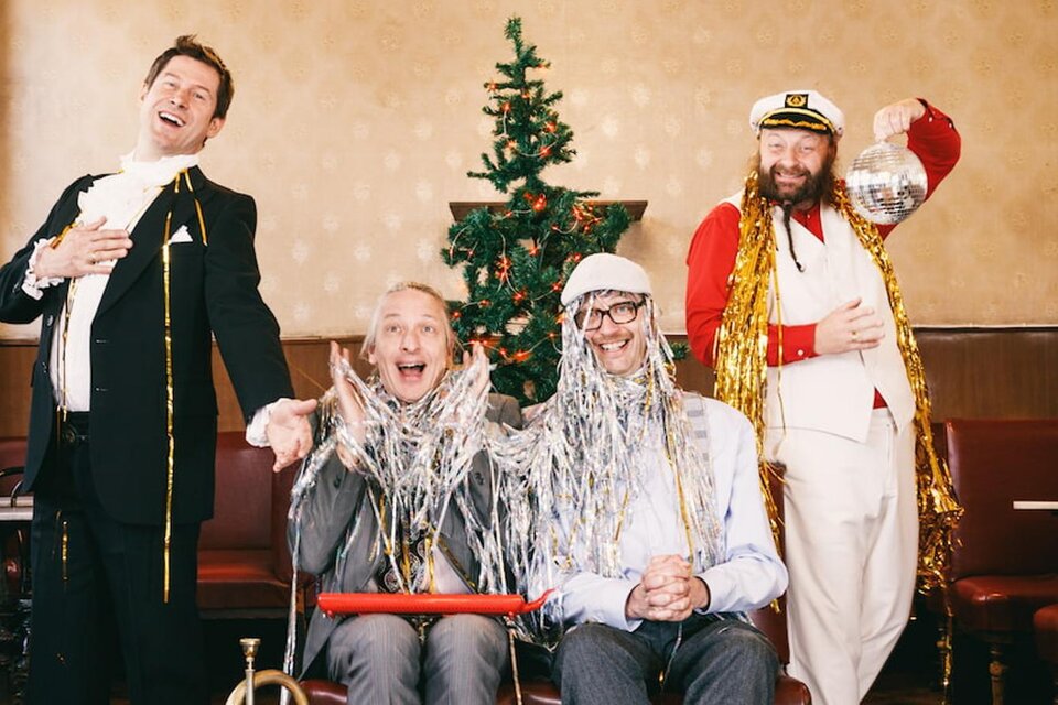 Vier Männer feiern eine fröhliche Weihnachtsstimmung. Sie tragen festliche Kleidung und stehen vor einem geschmückten Weihnachtsbaum. | © (c)Maria Frod