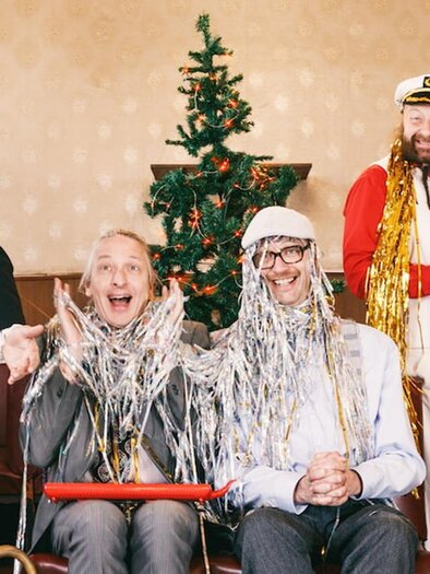 Vier Männer feiern eine fröhliche Weihnachtsstimmung. Sie tragen festliche Kleidung und stehen vor einem geschmückten Weihnachtsbaum. | © (c)Maria Frod