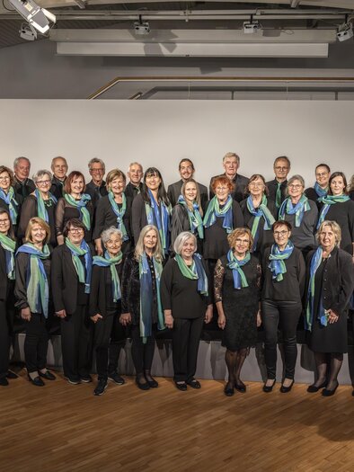A large choir is standing on a stage and smiling at the camera. The members are wearing black clothing with turquoise scarves. | © Ton.Art_Zurk