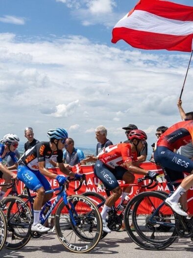 Eine Gruppe Radfahrer fährt auf einer Rennstrecke. Im Hintergrund schwenkt ein Zuschauer eine rot-weiß-rote Flagge. | © EXPA