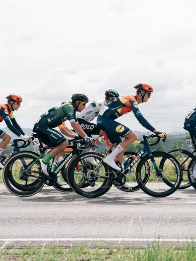A group of cyclists is racing on a road. The sky is cloudy and the landscape in the background is green. | © EXPA