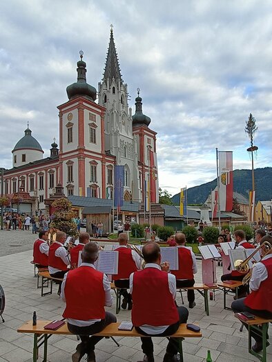 Eine Blaskapelle spielt vor einem beeindruckenden Gebäude auf dem Platz. Der Himmel ist bewölkt und die Atmosphäre ist festlich. | © Gabi Stebetak