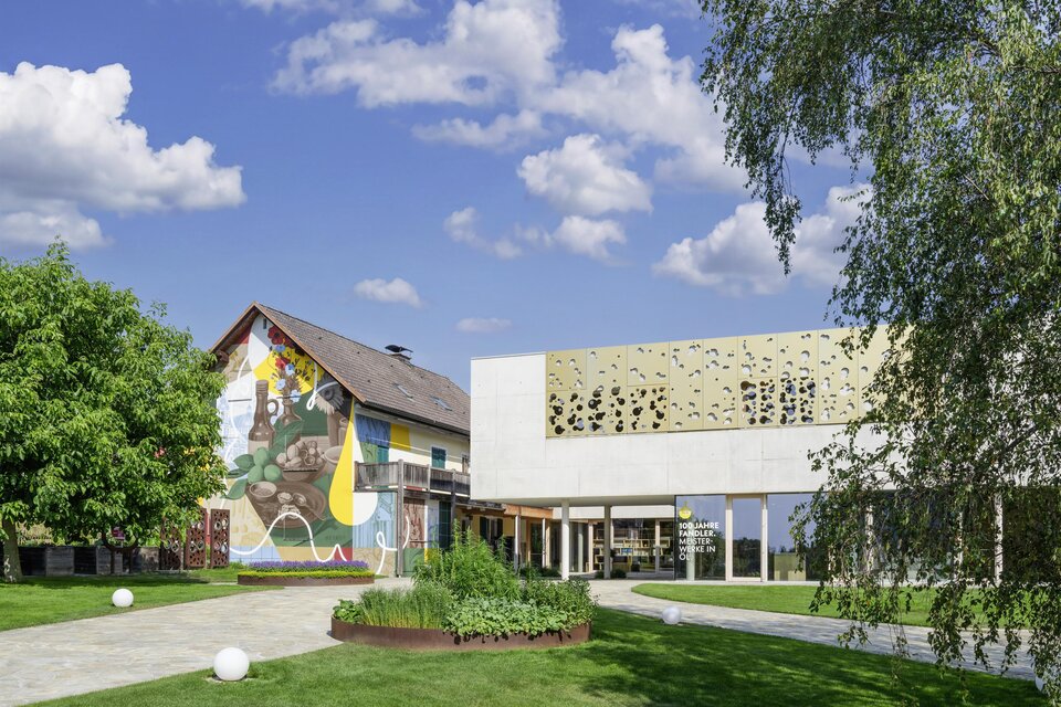 Ein modernes Gebäude neben einem traditionellen Haus mit buntem Wandgemälde. Die Landschaft ist grün mit Rasenflächen und Bäumen unter einem klaren Himmel. | © Peter Schmid