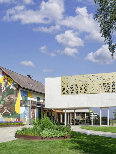 Ein modernes Gebäude neben einem traditionellen Haus mit buntem Wandgemälde. Die Landschaft ist grün mit Rasenflächen und Bäumen unter einem klaren Himmel. | © Peter Schmid