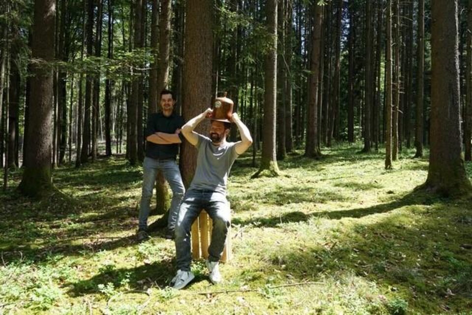 Zwei Männer stehen im Wald. Einer sitzt auf einem Hocker und trägt einen Hut. | © Destillerie Fuxbau OG