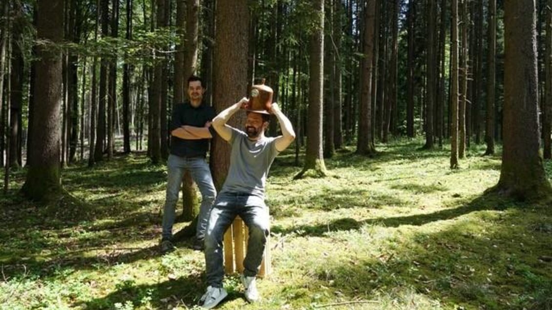 Zwei Männer stehen im Wald. Einer sitzt auf einem Hocker und trägt einen Hut. | © Destillerie Fuxbau OG