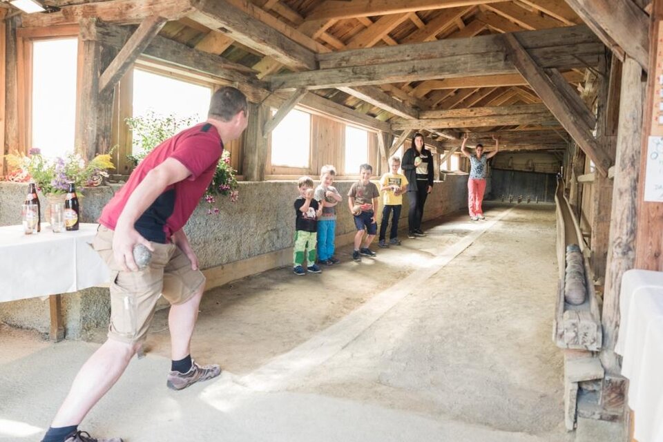 Eine Gruppe von Kindern steht in einer Scheune und beobachtet einen Mann, der einen Ball wirft. Die Atmosphäre ist freundlich und entspannt.