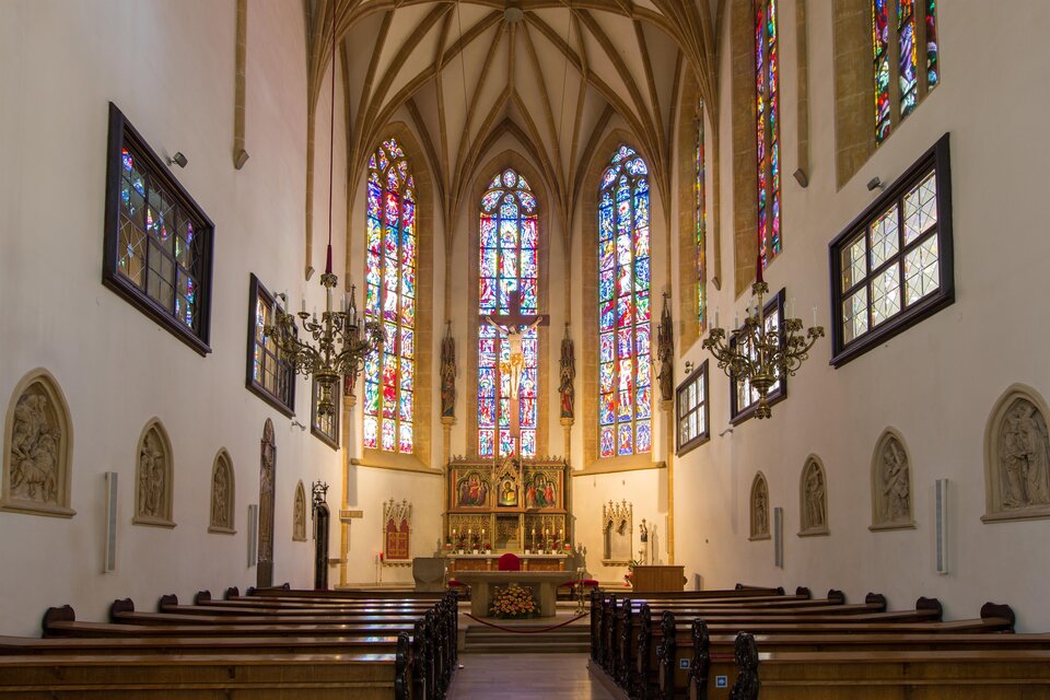 Eine ruhige Kircheninnenseite mit hohen gewölbten Decken und bunten Glasfenstern. Bänke stehen in geraden Reihen und der Altar ist im Vordergrund sichtbar.