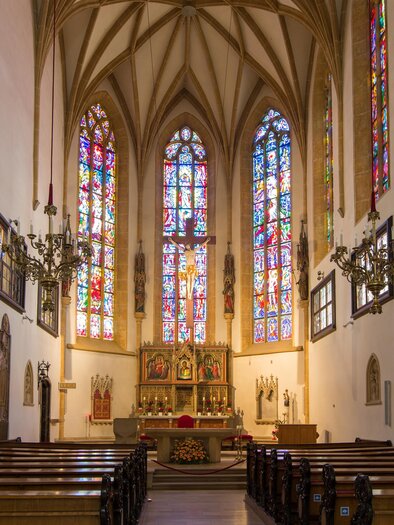 A quiet church interior with high vaulted ceilings and colorful stained glass windows. Benches are arranged in straight rows and the altar is visible in the foreground.