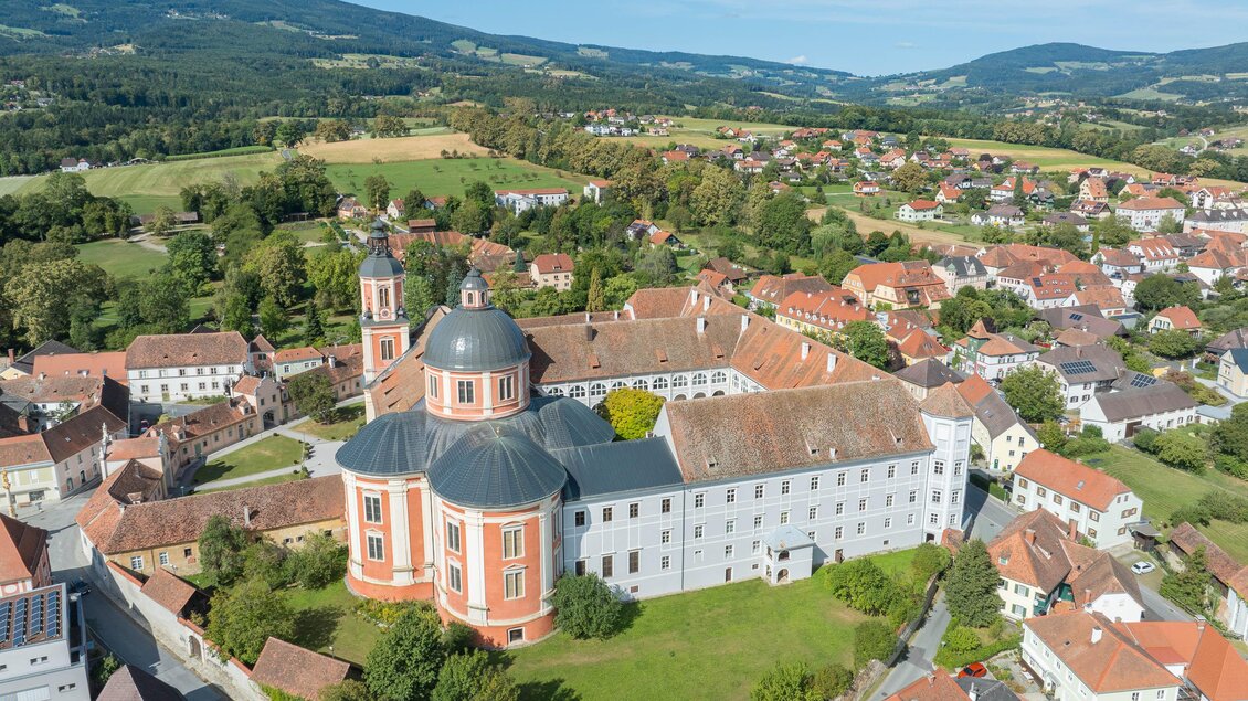 Eine große Klosteranlage mit einem gewölbten Dach und grünen Wiesen. Im Hintergrund sind sanfte Hügel und Wohnhäuser zu sehen. | © Helmut Schweighofer