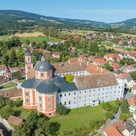A large monastery complex with a vaulted roof and green meadows. In the background, gentle hills and residential houses can be seen. | © Helmut Schweighofer