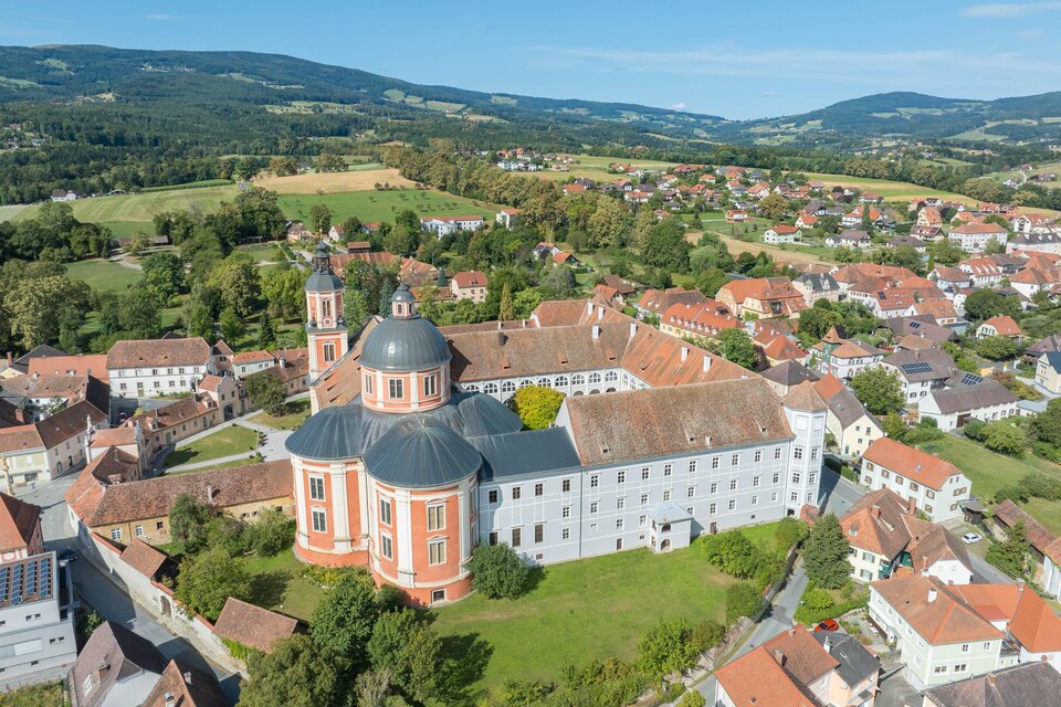 Eine große Klosteranlage mit einem gewölbten Dach und grünen Wiesen. Im Hintergrund sind sanfte Hügel und Wohnhäuser zu sehen. | © Helmut Schweighofer