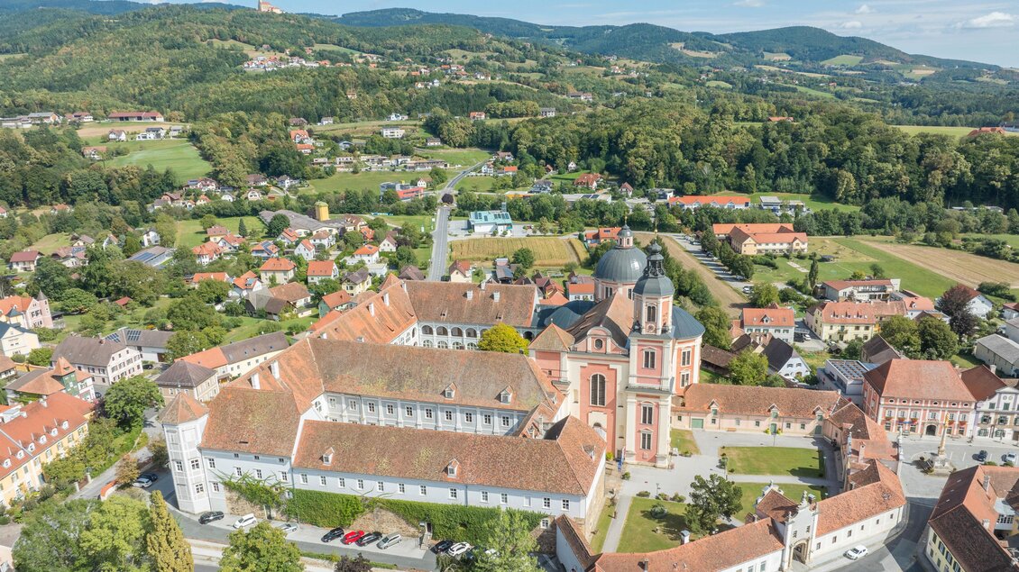 Eine beeindruckende Luftaufnahme eines historischen Klosters mit roten Dächern und einer markanten Kirche. Die umliegende Landschaft ist grün und hügelig. | © Helmut Schweighofer