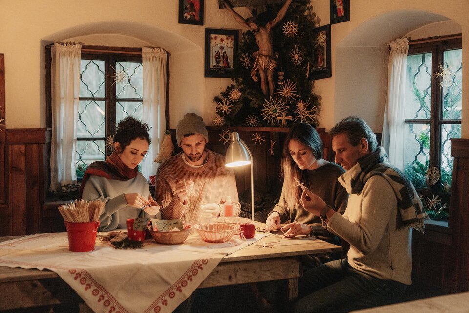 Eine Gruppe von vier Personen sitzt an einem Tisch und bastelt Weihnachtsdekoration. Im Hintergrund ist ein geschmückter Weihnachtsbaum zu sehen. | © (c) Region Graz - studio draussen