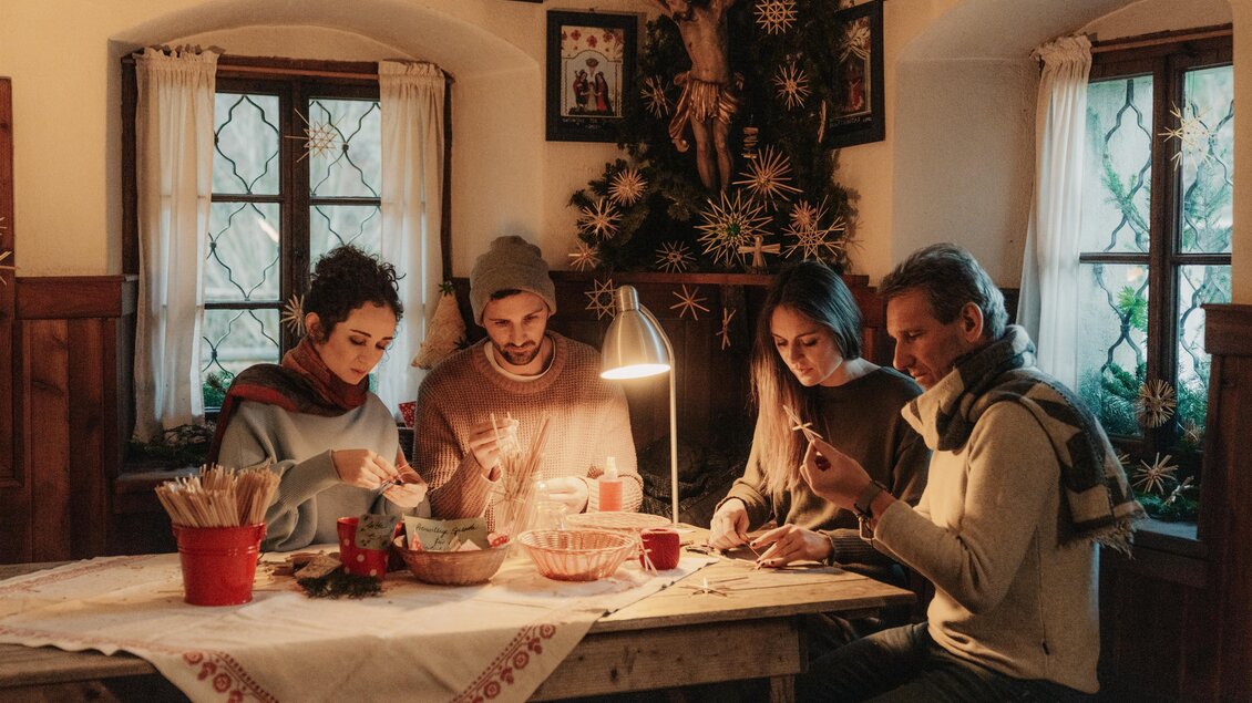 Eine Gruppe von vier Personen sitzt an einem Tisch und bastelt Weihnachtsdekoration. Im Hintergrund ist ein geschmückter Weihnachtsbaum zu sehen. | © (c) Region Graz - studio draussen