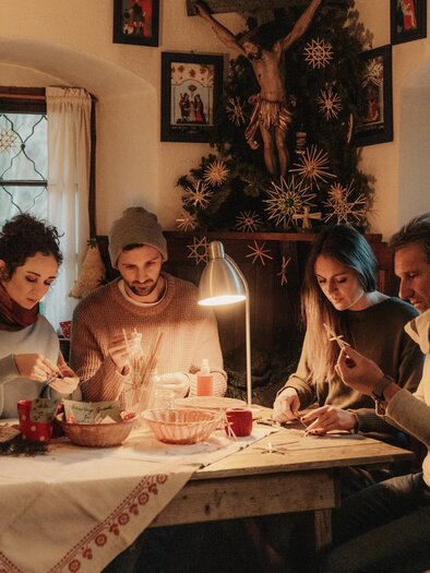 A group of four people is sitting at a table making Christmas decorations. In the background, a decorated Christmas tree can be seen. | © (c) Region Graz - studio draussen