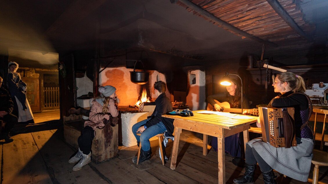 Eine gemütliche Stube mit mehreren Personen, die um ein Feuer sitzen. Eine Frau spielt ein Musikinstrument, während andere zuhören. | © Region Graz - Harry Schiffer