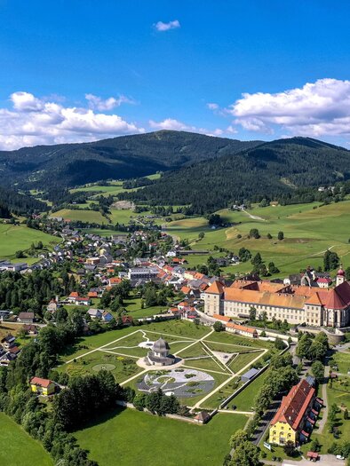 Eine malerische Landschaft mit einem historischen Gebäude und sanften Hügeln. Im Hintergrund sind grüne Wiesen und ein blauer Himmel zu sehen.