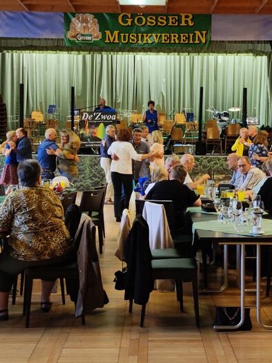 People are dancing and having fun in a festive hall with tables. In the background, a band is visible on stage. | © Referat Soziales, Familien & Frauen