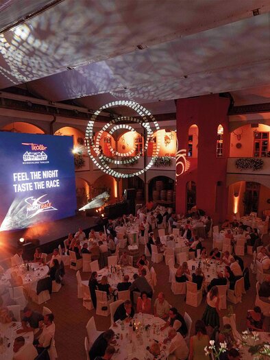 A festive event in an elegantly decorated hall. Tables are set, and a large screen displays a presentation. | © Rene Strasser