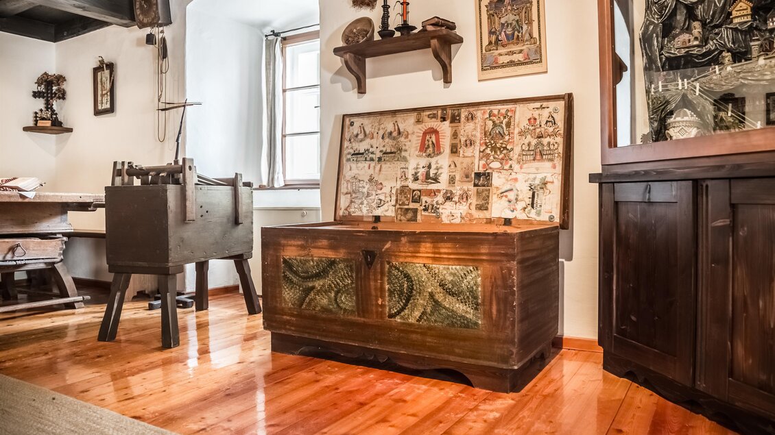 Ein gemütlicher, traditioneller Raum mit Holzfußboden und alten Möbeln. Im Vordergrund steht eine kunstvoll verzierte Truhe. | © Fred Lindmoser