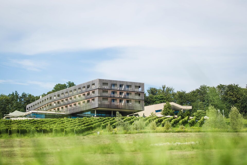 Ein modernes Hotel umgeben von Weinbergen und grüner Landschaft. Im Hintergrund sind sanfte Hügel und ein klarer Himmel zu sehen. | © LOISIUM