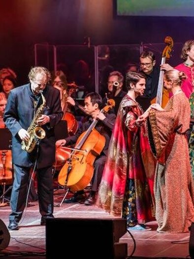 A lively stage performance with musicians and dancers. In the background, an orchestra plays while the artists wear colorful costumes. | © HC_KoenigderLoewen_Bild
