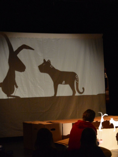 Eine Puppenspielaufführung mit Schatten von einem Hasen und einer Katze auf einer Leinwand. Im Vordergrund sind die Spieler mit Requisiten zu sehen. | © Theater Bredouille